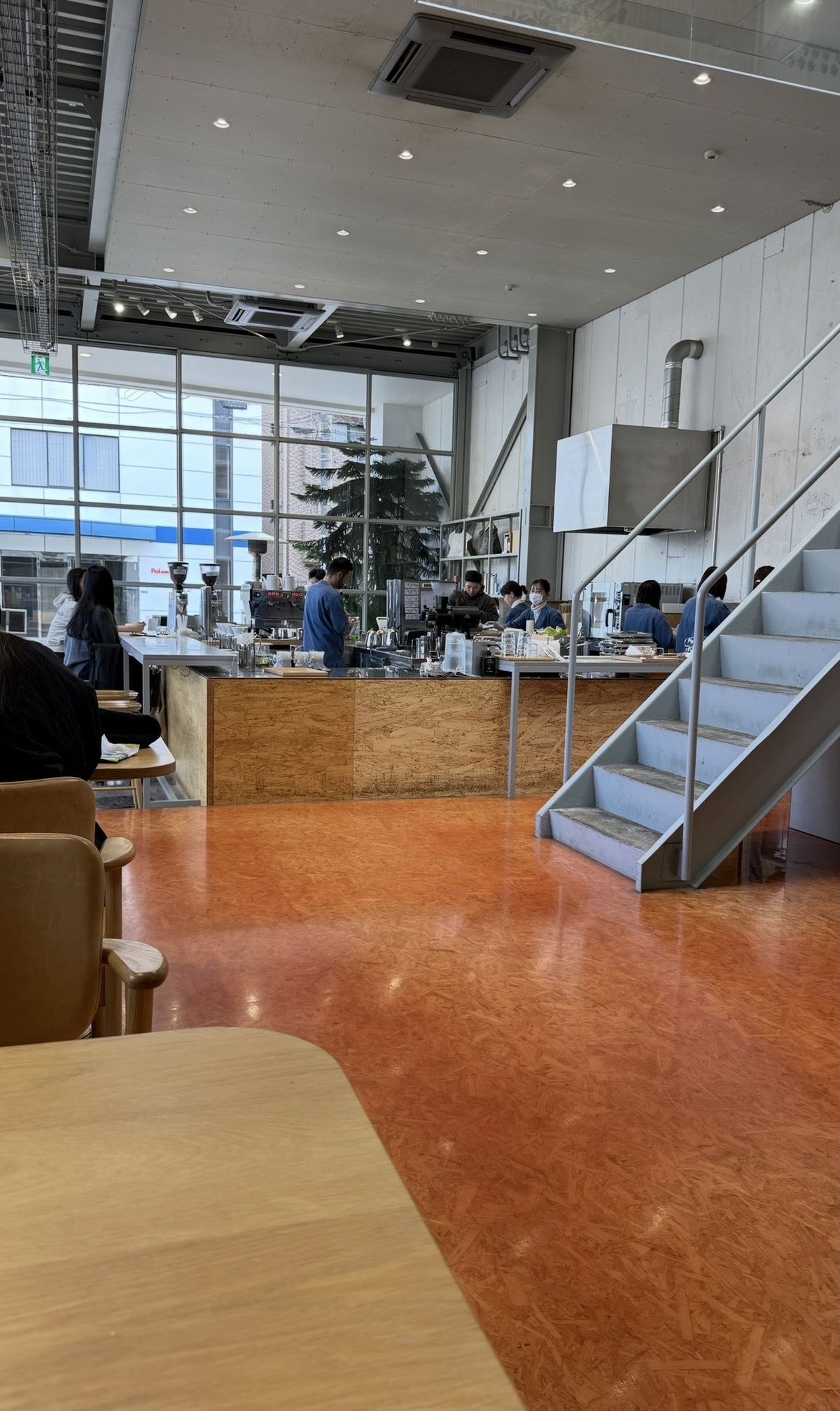 Interior de un café amplio con gente en la barra y mesas, piso naranja y escalera metálica